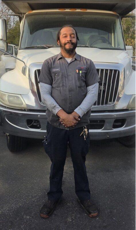 Full body photo of Michael Blanding, Diagnostic Specialist, standing in front of the San Antonio shop, ready to perform DOT-compliant inspections and Mobile A/C (MVAC) refrigerant services.